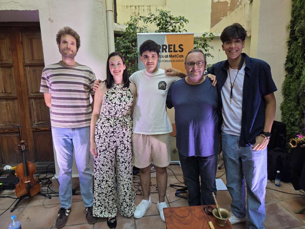 Five people stand in a row outdoors, smiling for a group photo with a banner behind them and musical instruments nearby.