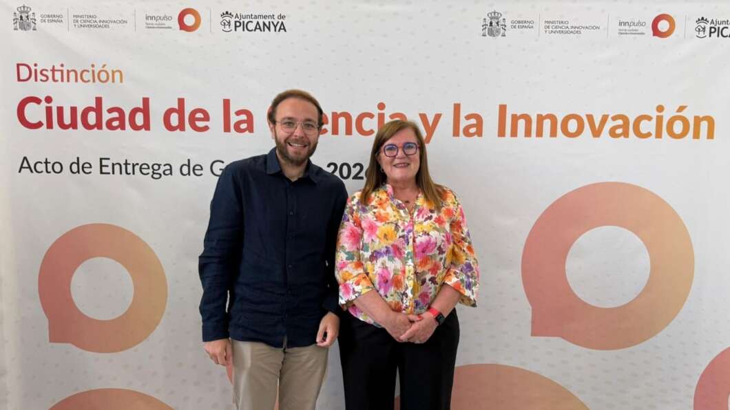 Man and woman posing in front of a banner that says 'Distinción Ciudad de la Ciencia y la Innovación' with logos at the top.