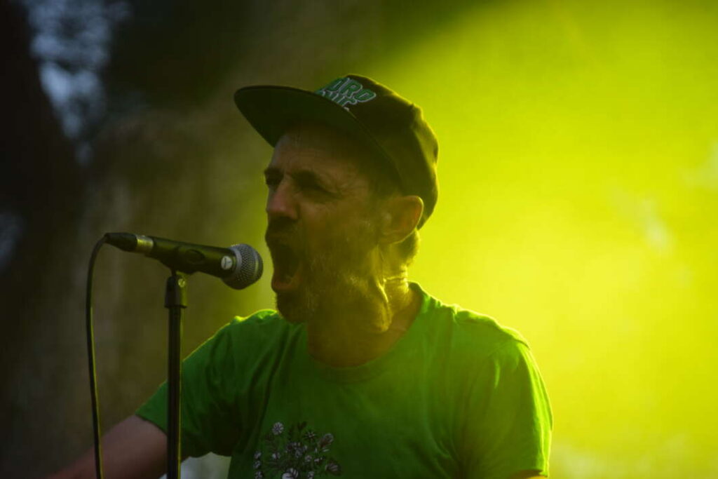 Male singer in a green shirt and cap performs into a microphone on stage with bright yellow lighting in the background.