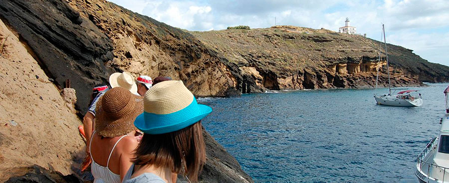 People wearing hats stand on rocky shore overlooking blue sea with a sailboat and a distant lighthouse.
