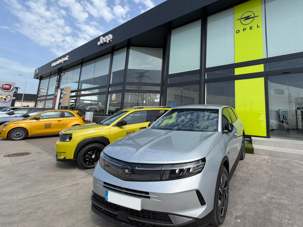 Silver SUV in the foreground of a glass-walled car showroom with bright yellow Opel branding and a row of yellow cars nearby.