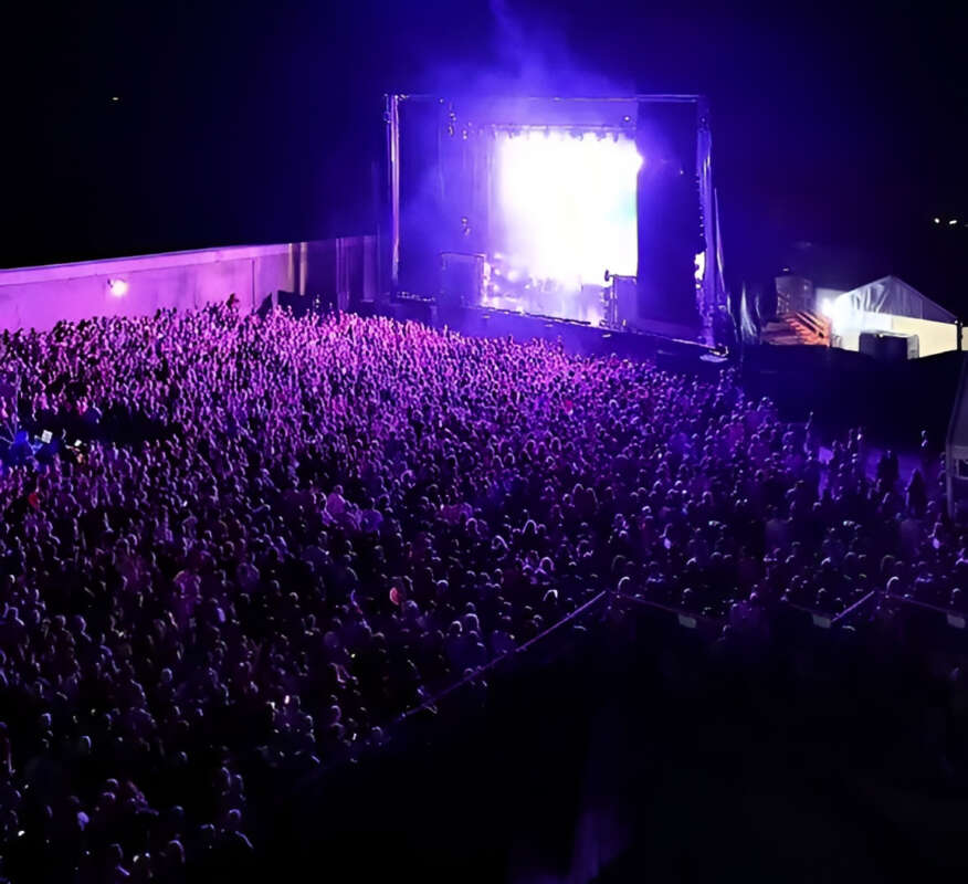 Large outdoor concert crowd at night facing a brightly lit stage with purple lights and a big screen.