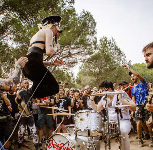 Musician in a black outfit leaps over a silver drum kit as a crowded outdoor audience watches a daytime show.