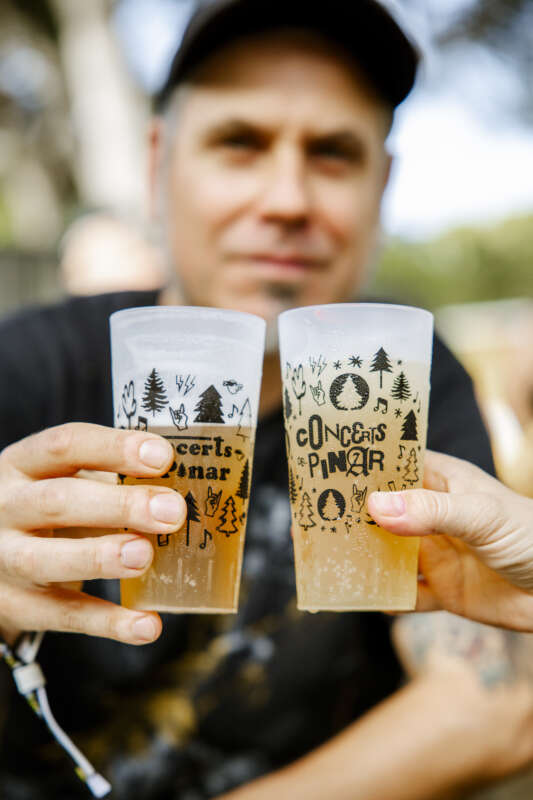 Two frosty beer cups decorated with trees and concert graphics are raised for a toast, held by a person in a cap in a sunny outdoor setting.