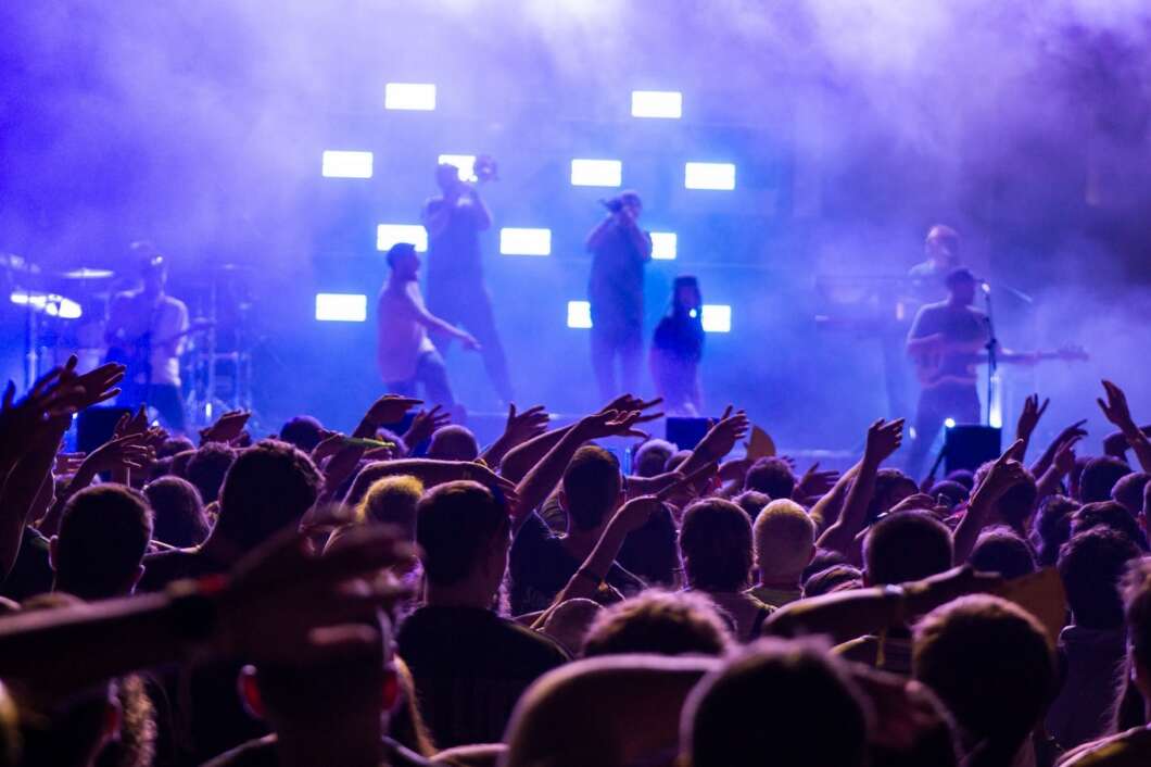 Crowd at a live concert with hands raised, performers on a smoky blue-lit stage in the background.