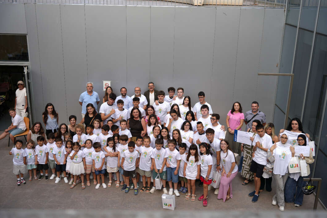 Large group of children and adults posing together for a group photo outside a modern building, many wearing white T-shirts with a small logo on the front.