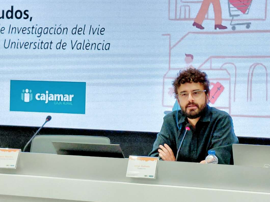 Bearded man wearing glasses sits at a conference panel with a microphone, in front of a screen displaying the Cajamar logo and light graphics.