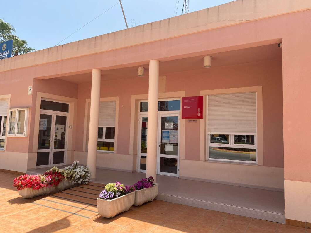 Front of a pink municipal building with two pillars, glass doors, and colorful flower planters outside the entrance.