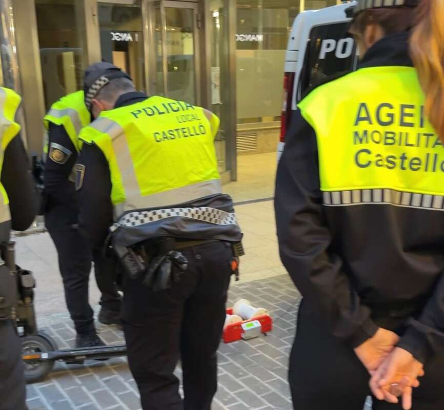 Policías locales de Castellón con chalecos reflectantes amarillos en una acera peatonal, frente a una tienda, junto a un scooter eléctrico y una caja roja en el suelo.