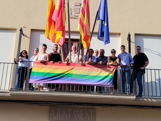 Diverse group of people on a balcony holding a large rainbow Pride flag, with Catalan and blue flags behind them near a municipal building.