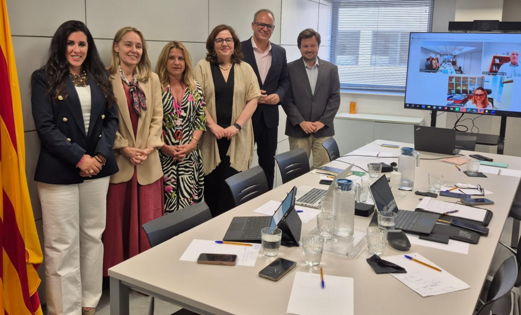 Six professionals pose for a photo in a modern meeting room, a long table with laptops, papers, and glasses in view, and a video conference screen on the right showing remote participants.
