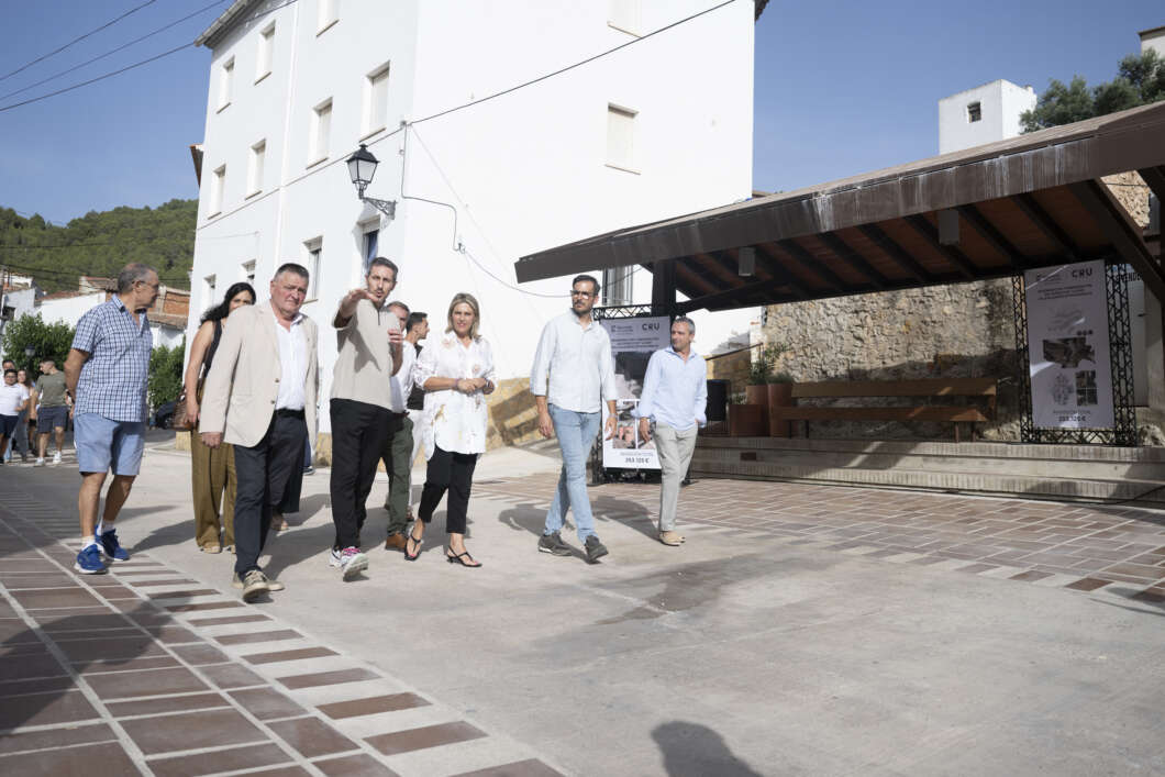 Group of people walking together in a sunny plaza beside a white building with a covered stage nearby.