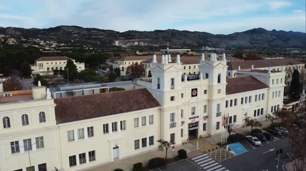 Aerial view of a pale-yellow/white institutional building with twin towers, flags, and a crosswalk in front, set against hills in the background.