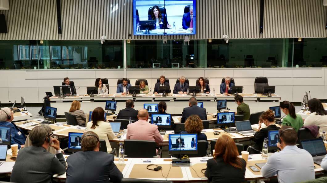 Panel of officials seated at a long desk in a modern conference room, attendees using laptops and screens throughout the room.