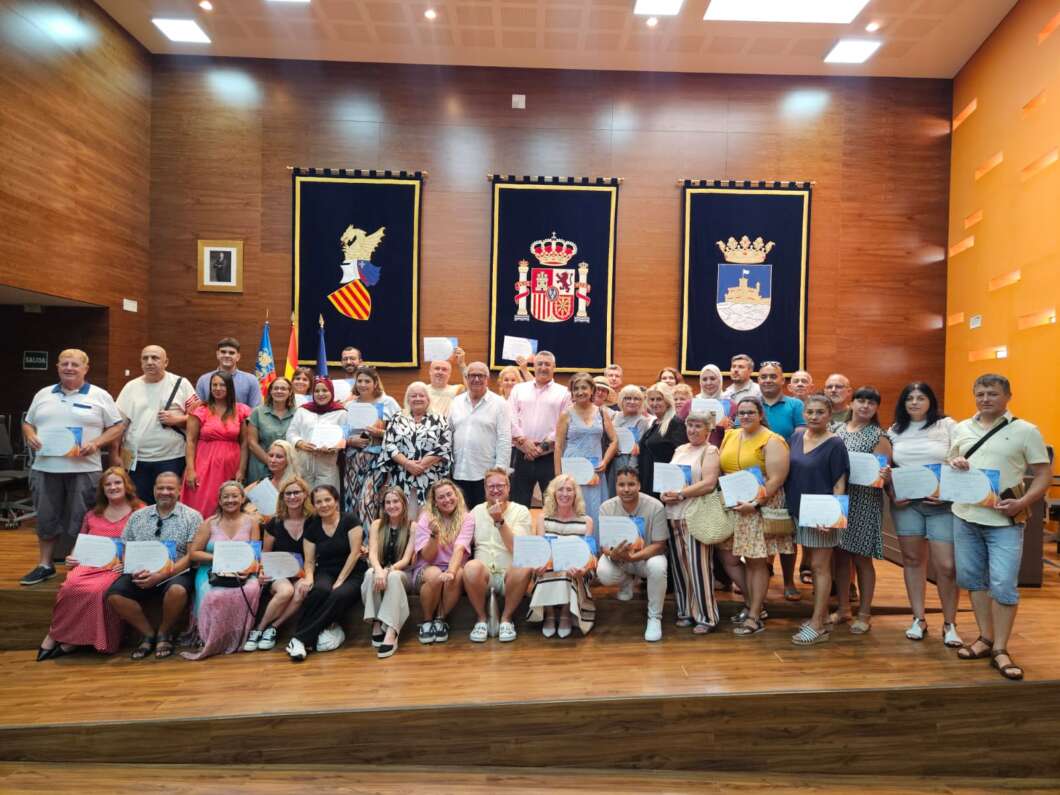 Group of people on a stage holding certificates after an awards ceremony in a wood-paneled hall with flags and banners behind them, smiling for the photo.
