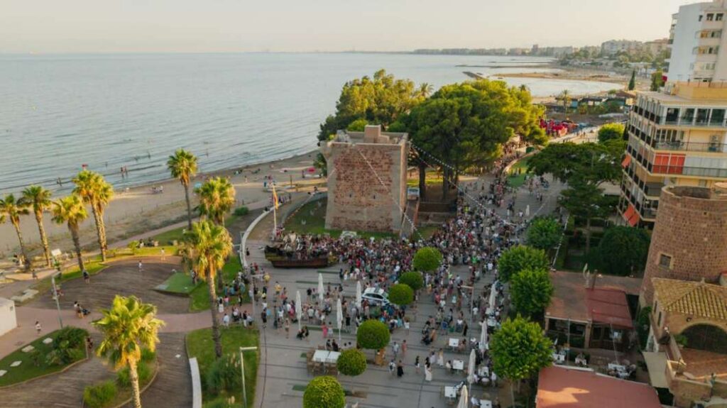 Aerial view of a seaside plaza with a crowd, palm trees, and a stone fortress near the beach and ocean.