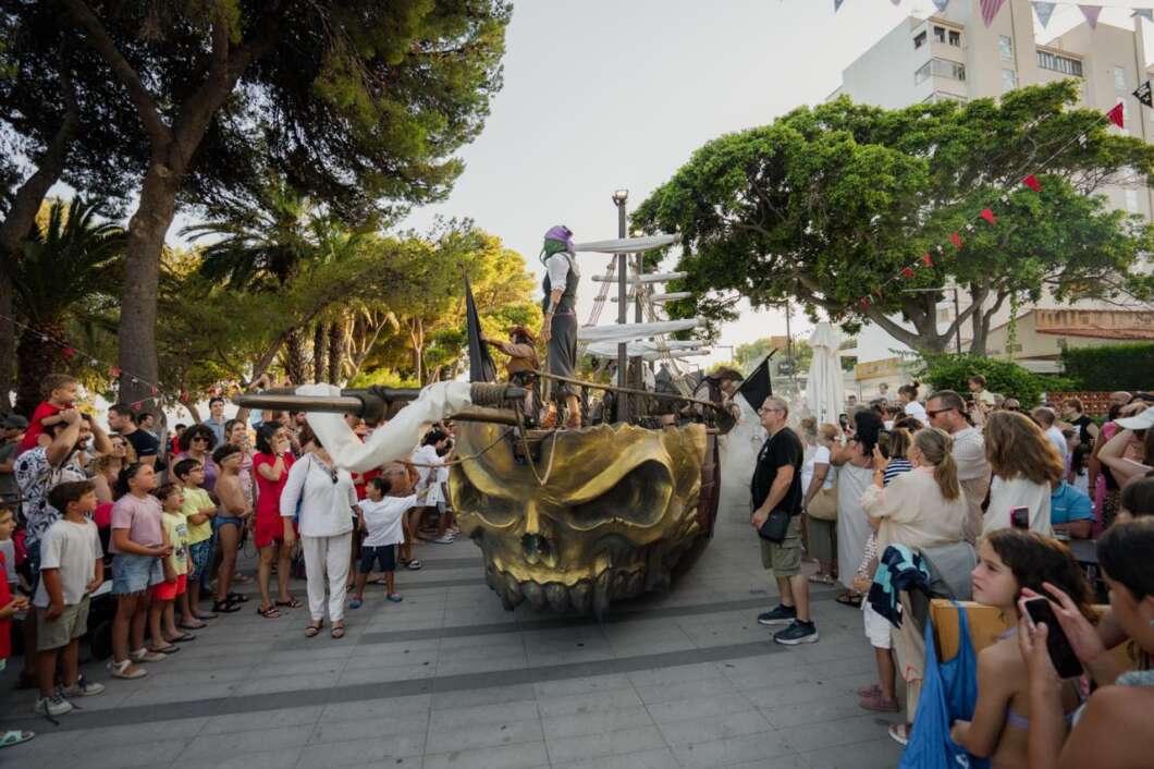 Crowd watches a giant golden skull with a ship perched on top during a street parade in a sunny plaza.