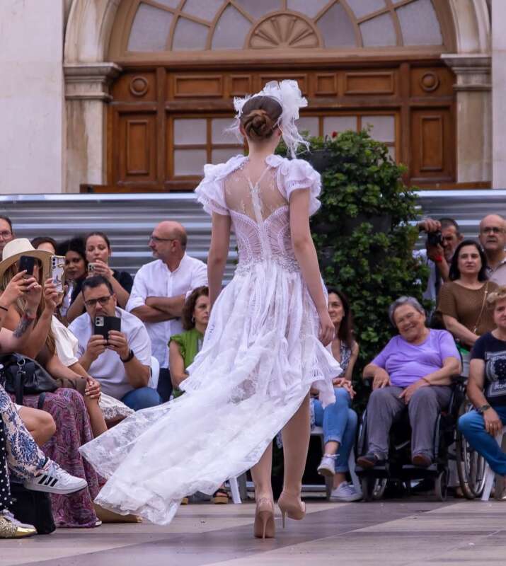 Back view of a model in a white lace dress walking down a runway as spectators photograph her.