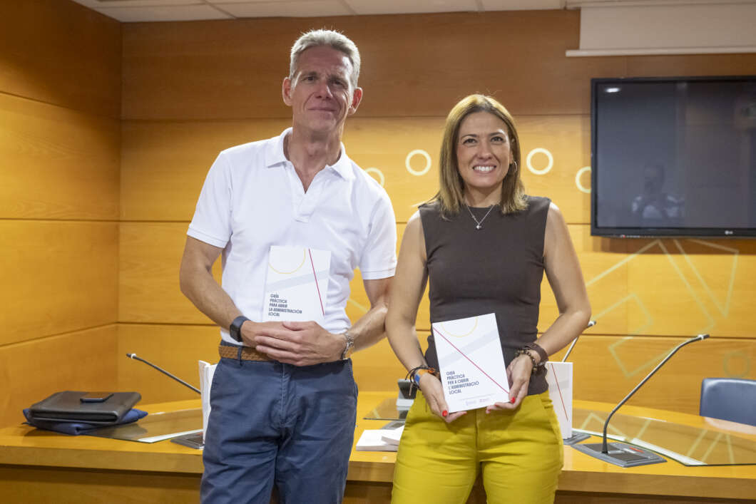 Two adults stand in a wood-paneled conference room, each holding a booklet and smiling for the camera.