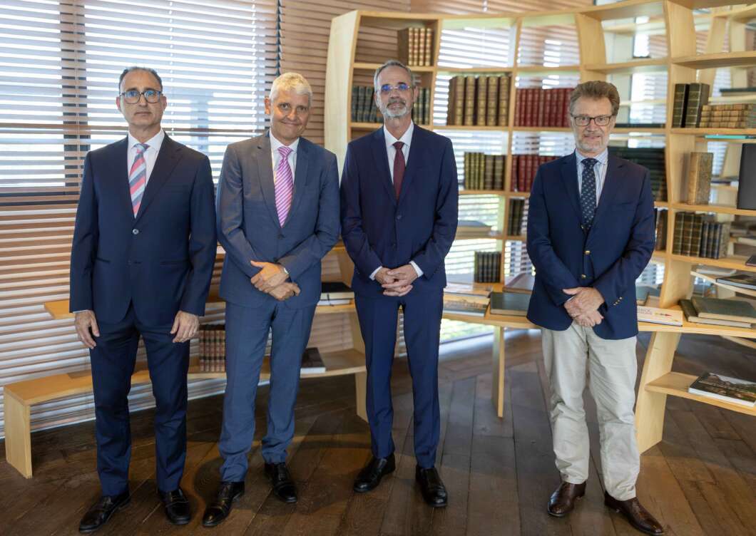 Four men in suits stand side by side in a modern library with wooden shelves full of books behind them, posing for a photo.