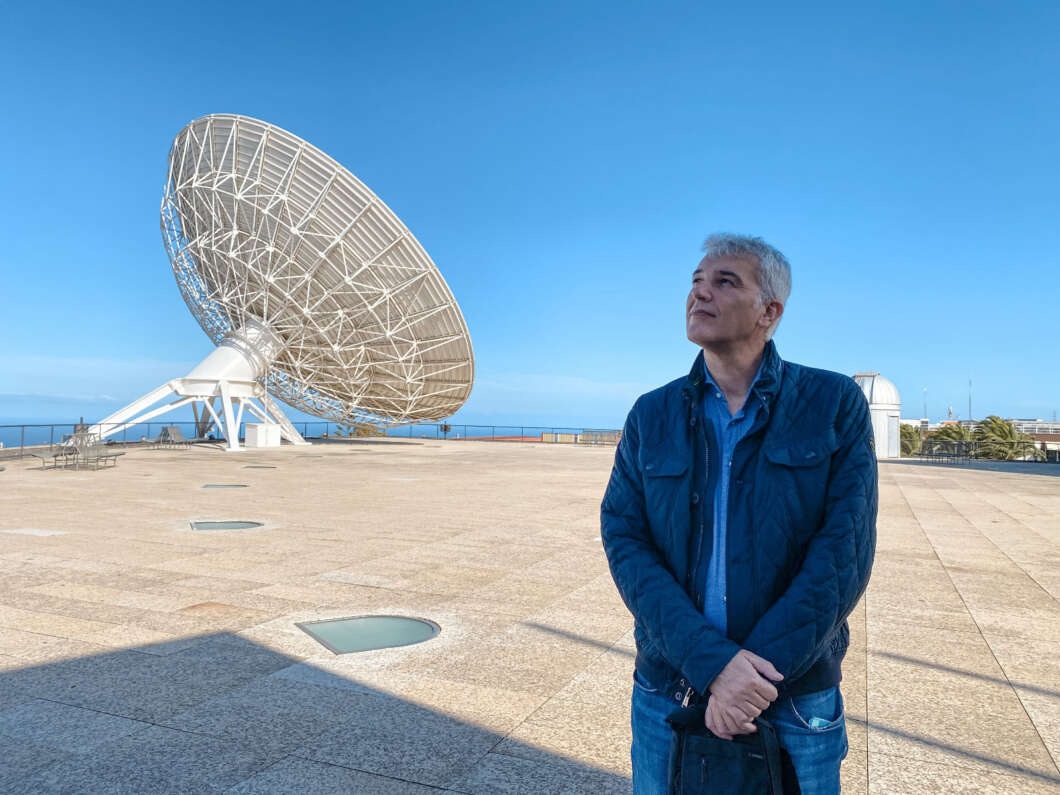 Man standing on a sunny rooftop next to a large satellite dish, looking up at the sky with the ocean visible in the distance.