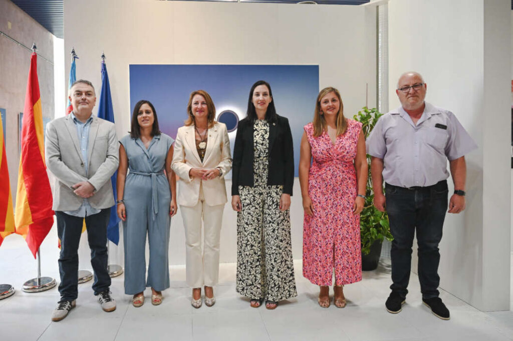 Six adults in smart-casual to business attire pose for a group photo indoors, with flags on the left and a blue backdrop behind them.