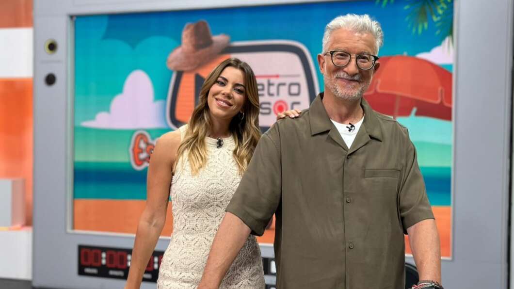 Two smiling TV hosts pose together in a studio, a woman in a white lace dress and a man in a green shirt, with a colorful retro set behind them.