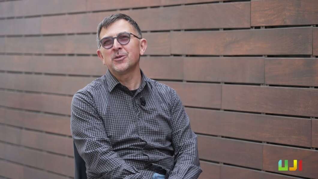 Medium shot of a man in a checkered shirt and glasses seated in front of a brown wooden fence, with a lapel mic clipped to his chest.