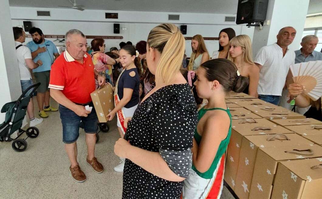 A volunteer in a red shirt hands a boxed gift to a girl while people wait in line; many cardboard boxes are stacked on a table nearby.