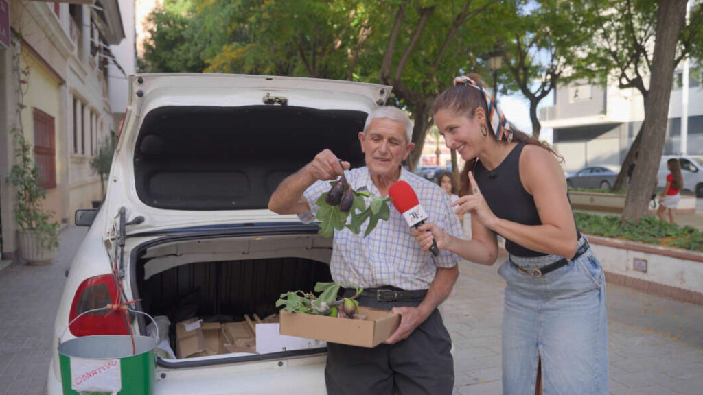 Elderly man shows a potted plant to a reporter beside an open car trunk filled with boxes on a city street.