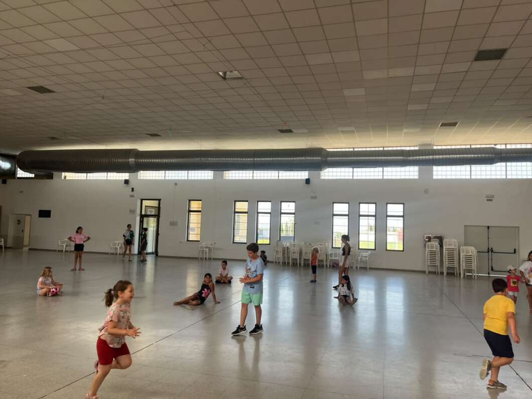 Children playing and running in a bright, spacious indoor gym with large windows and stacked chairs along the wall.