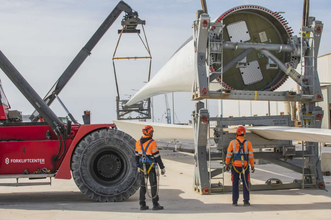 Two workers in orange safety gear stand on a concrete surface watching a large wind turbine blade being moved by a red forklift and hoisting rig.