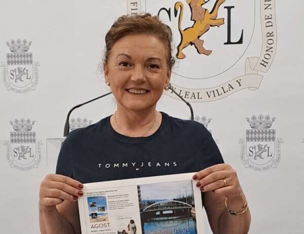 Smiling woman in a navy Tommy Jeans shirt holds a printed brochure in front of a backdrop featuring crests and emblems.