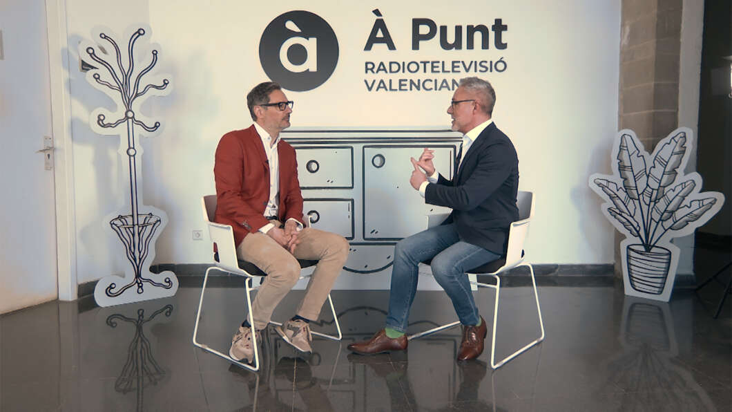 Two men sit on white chairs facing each other in a TV studio, mid-conversation, with an À Punt Radiotelevisió Valenciana backdrop behind them.