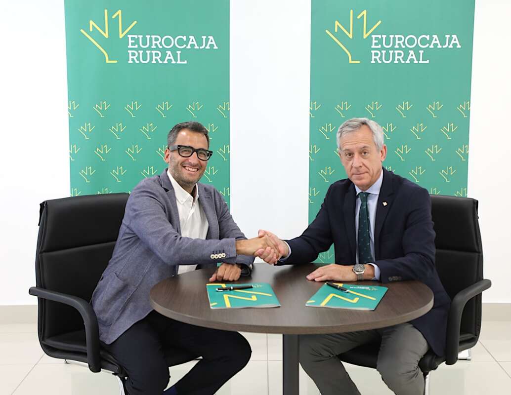Two men in business attire shake hands over a round table, with Eurocaja Rural banners in the background.