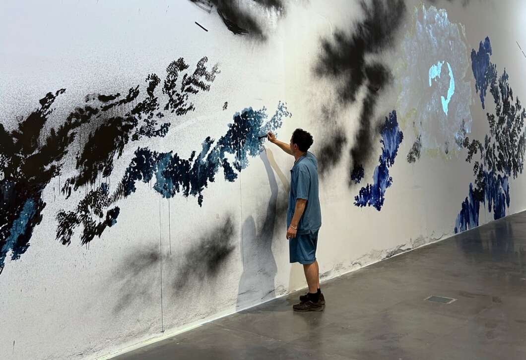 A man in blue clothing paints a long abstract mural with blue and black brushstrokes on a white wall.