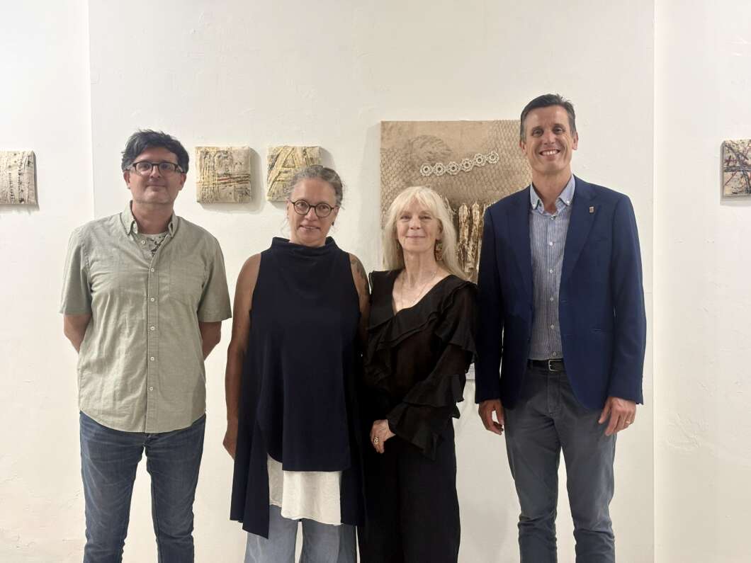 Four adults stand side by side in a bright white-walled art gallery, smiling with textured artworks on the wall behind them.