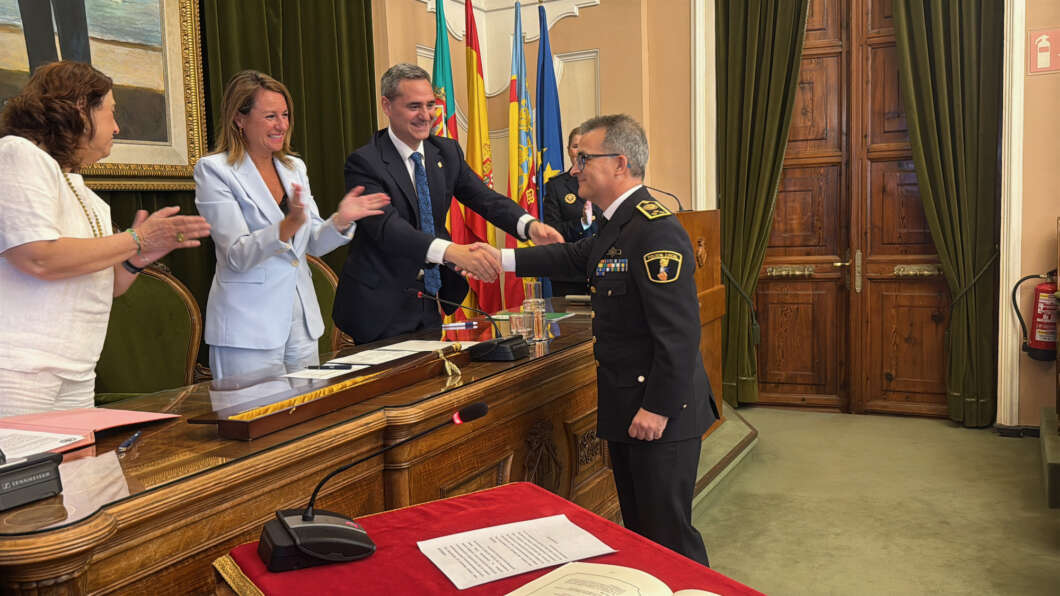 Man in a suit shakes hands with a uniformed officer across a ceremonial table as officials applaud in the background with flags behind them.