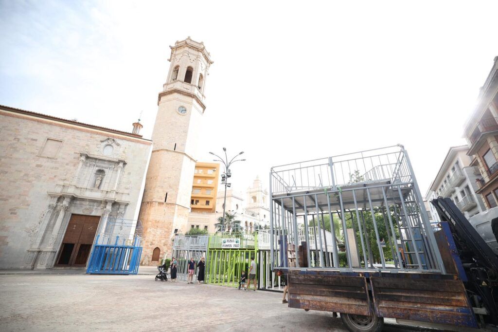 Historic plaza with a tall bell tower and ornate stone church; a metal cage-like structure sits on a truck in the foreground.