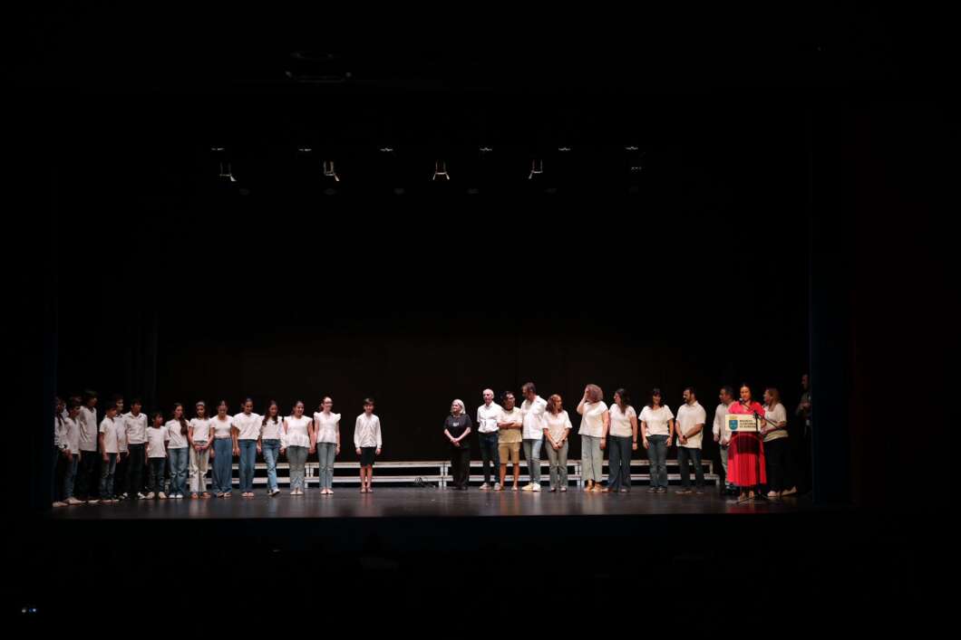 Group of people on a dark theater stage, many in white shirts, standing in a line for a ceremony or performance.