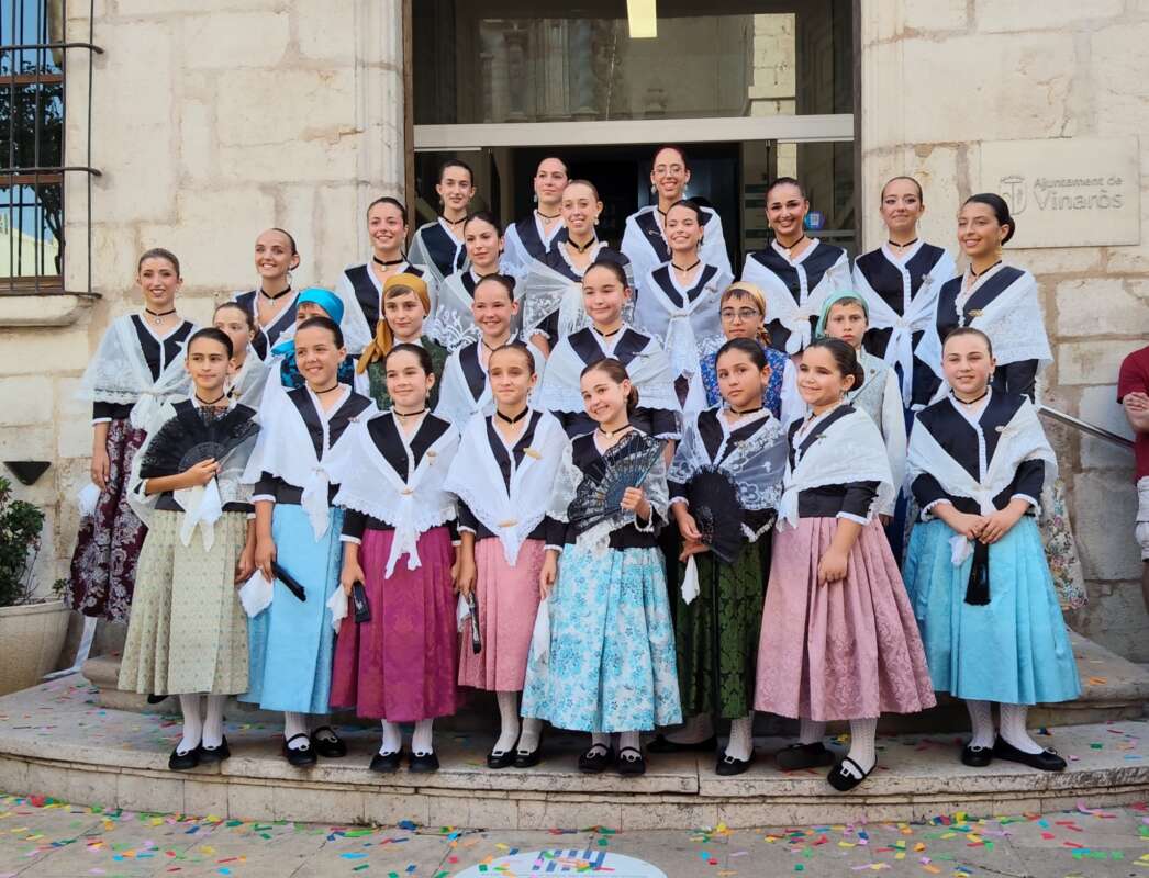 Group of girls in traditional folk costumes posing on steps outside a stone building for a group photo.