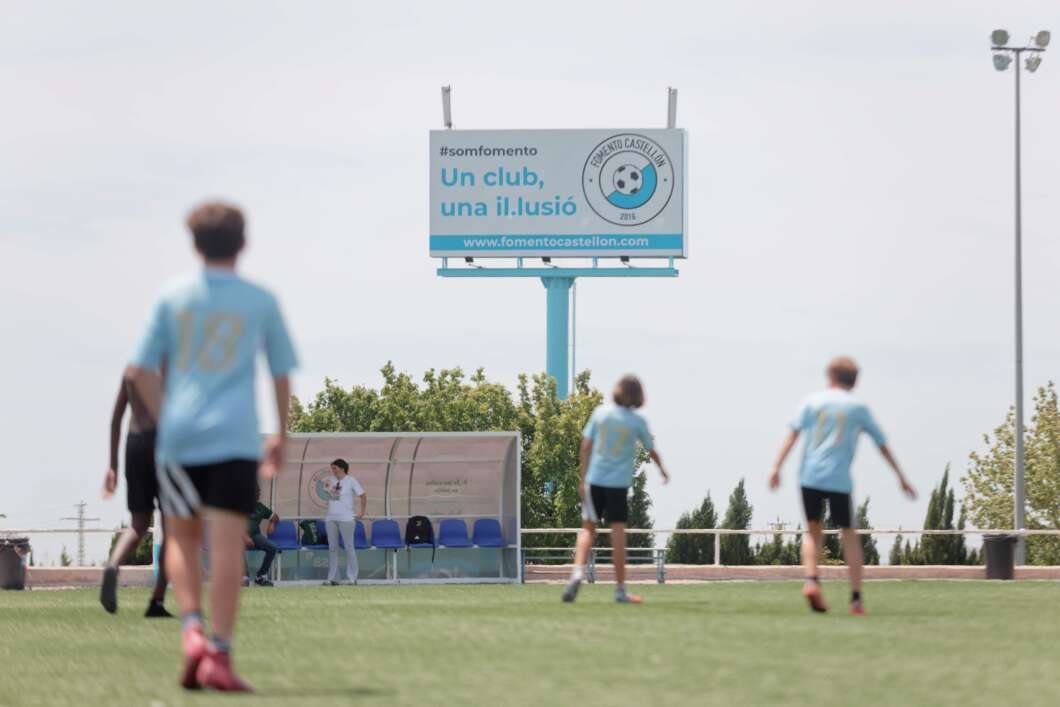 Soccer field with players in light blue jerseys and a bench shelter in the background, billboard behind them.