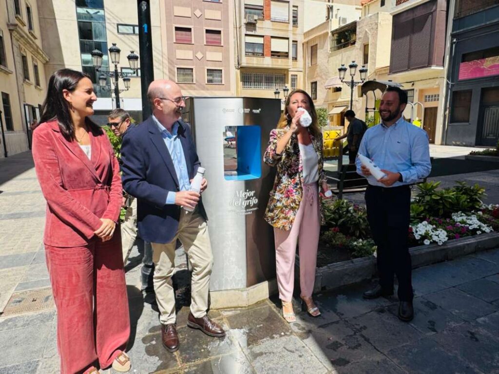 Group of four adults standing around a stainless water dispenser in a sunny plaza, one woman drinking from a bottle and others smiling and chatting.