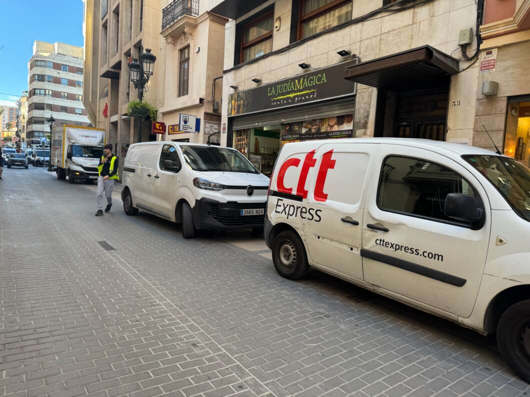 Street with white delivery vans parked along a pedestrian street; a worker in a safety vest stands nearby.