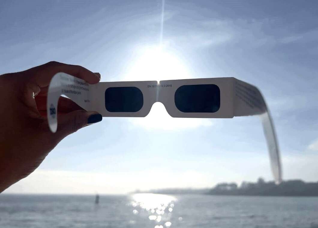 Hand holding white cardboard 3D glasses with blue lenses against the sun over the ocean.