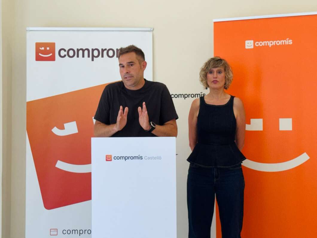 Man at a podium giving a speech, with a woman standing beside him, at a compromís Castelló event with orange branding in the background.