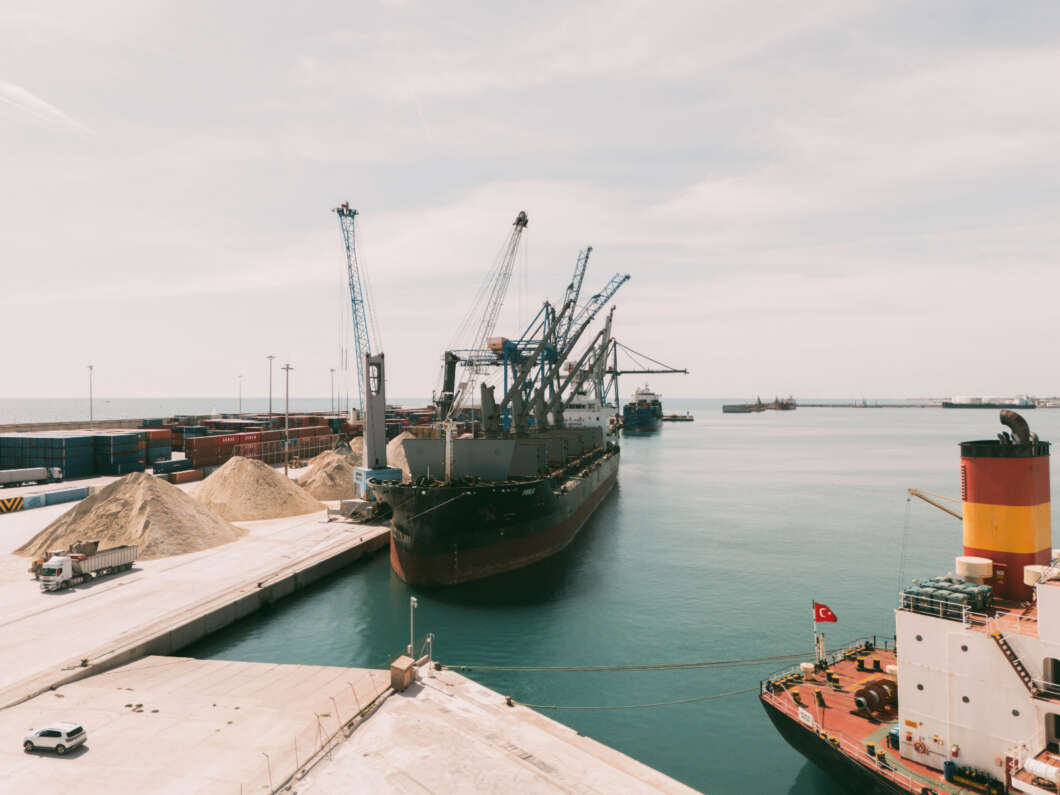 Port scene with a large cargo ship and cranes unloading at a quay, stacked containers and sand piles nearby by calm turquoise water, Turkey flag on a nearby vessel.
