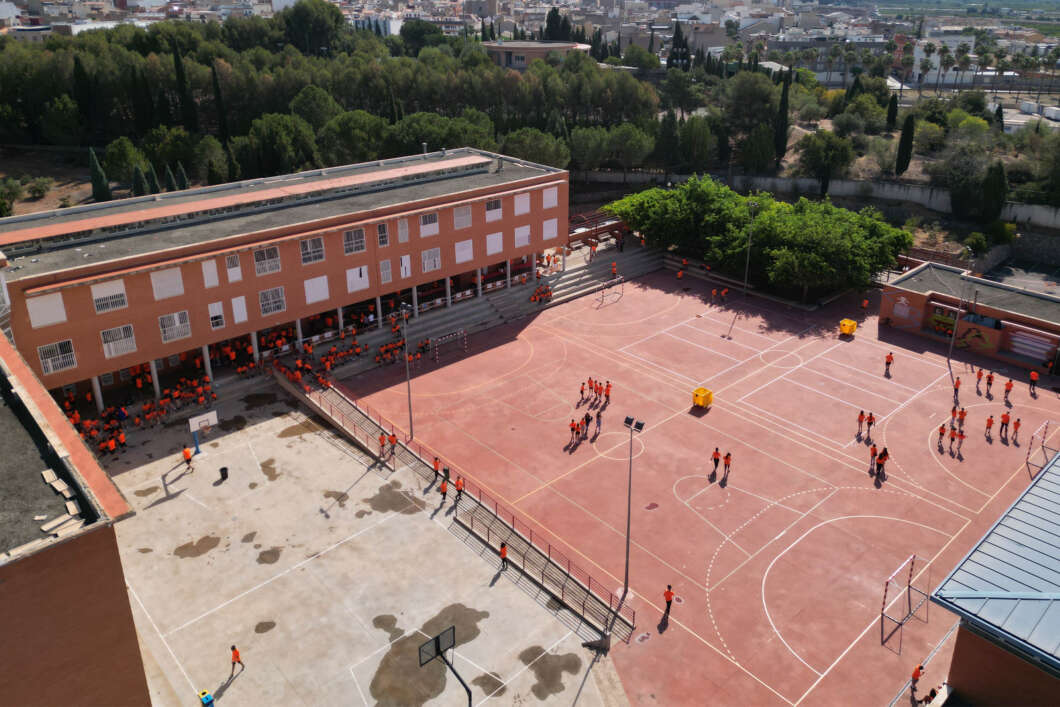 Aerial view of a school courtyard with a red basketball/handball court, students in orange uniforms, and a long brick building nearby.