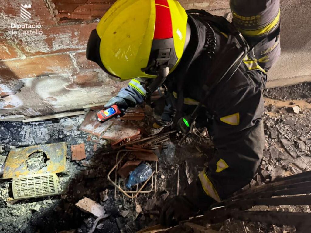 Firefighter in a yellow helmet and protective suit searches through rubble against a brick wall, with a municipal logo visible in the corner.