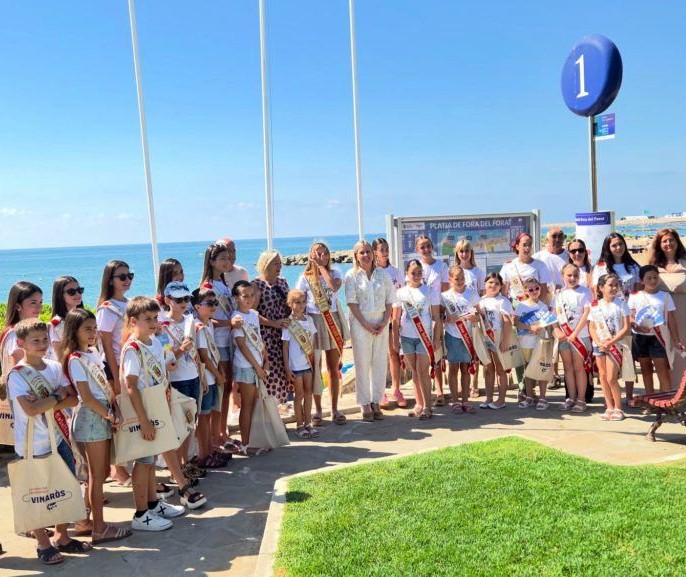 Group of children and adults posing on a seaside boardwalk for a photo, many wearing white shirts and sashes, with the ocean in the background and a large blue number-1 balloon overhead.